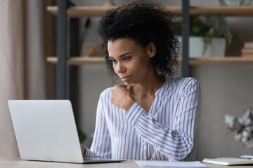 Thoughtful interested young african ethnicity business lady looking at laptop screen, working on online project, considering problem solution, web surfing information, preparing report or presentation
