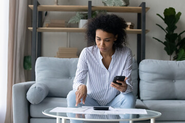 Concentrated young african american biracial woman holding telephone in hands, checking utility...