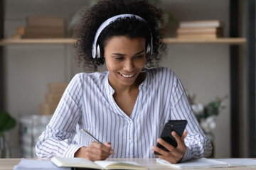 Smiling young african american woman in headphones holding telephone in hands, listening lecture or webinar online, writing notes in copybook, e-learning process, knowledge improvement concept.