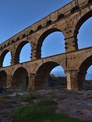 Fototapeta premium View of ancient Roman aqueduct Pont du Gard with majestic stone pillars above Gardon river after sunset near Vers-Pont-du-Gard, Occitanie, France.