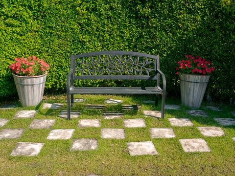 Empty black iron bench decorated between two flower pots on the green grass, the checkered pattern on bush background in the outdoor garden.