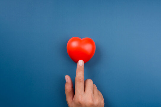 The Red heart ball is pointing or touching by finger on blue background. Love, care, sharing, giving, well-being, and donation concepts, minimal style.