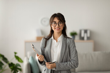 Portrait of cheerful female psychologist posing with clipboard, looking and smiling at camera, working in modern office - obrazy, fototapety, plakaty