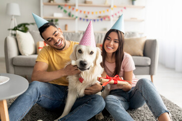 Millennial multiracial couple in party hats holding gift box, celebrating pet dog's birthday at home