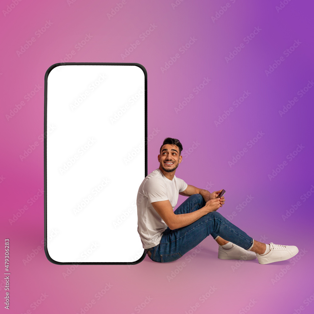 Poster young arab guy using cellphone while sitting near giant mobile phone with empty white screen, neon l