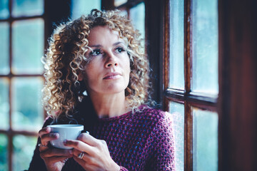Portrait of thoughtful attractive adult woman at home holding a cup of tea and looking outside the...