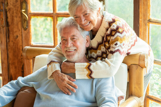 Happy and enjoyed senior caucasian couple smile and hug each other sitting on a chair in cozy home cabin chalet - two man and woman old elderly people lifestyle enjoying relax and relationship