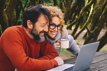 Happy modern couple use together laptop computer outdoors with trees and woods in background -...