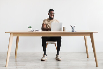 Happy young black man using laptop computer for online work at table in home office, free space
