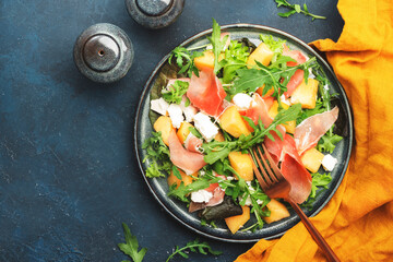 Summer salad with cantaloupe melon, prosciutto, white cheese and arugula on blue table background, top view, copy space