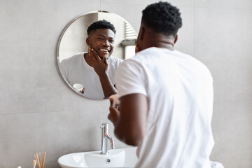 Happy black guy applying moisturizer cream on face in bathroom