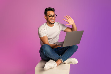 Young Arab guy sitting cross legged with laptop, waving at webcam, communicating to friend or colleague in neon light
