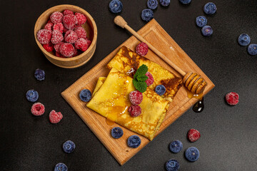 Delicious thin pancakes for breakfast on kitchen board with a bowl of raspberries, blueberries, fresh mint leaves and honey on dark background, top view