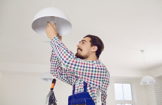 Electrician Changing Light Bulb. Young Man In Overall Work Uniform Standing On Ladder In Residential Building, Office, School Or Hospital And Screwing LED Lightbulb Into Modern White Ceiling Lamp