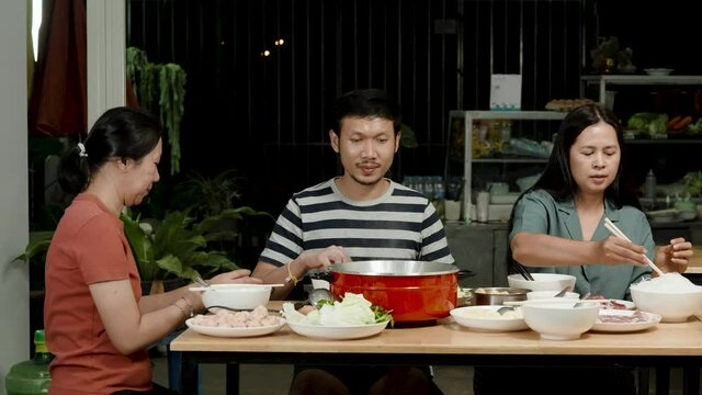 4k, A group of 3 Asian friends are eating sukiyaki in a restaurant in front of them with sukiyaki condiments and red sukiyaki pots.