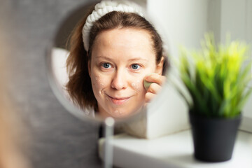 Mature woman does makeup at home in front of the mirror, applies BB cream with a sponge
