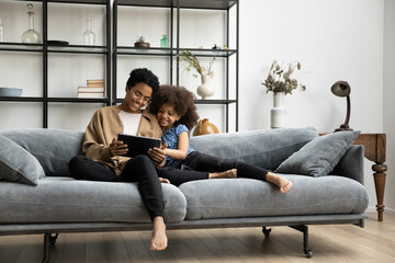 Happy Black mom and daughter kid using digital gadget for entertainment, relaxing on comfortable couch, watching movie, online series on tablet, talking on video call, smiling, laughing. Wide shot