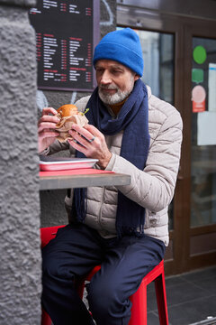 Senior male person eating his meal while sitting at the table with big tasty sandwich