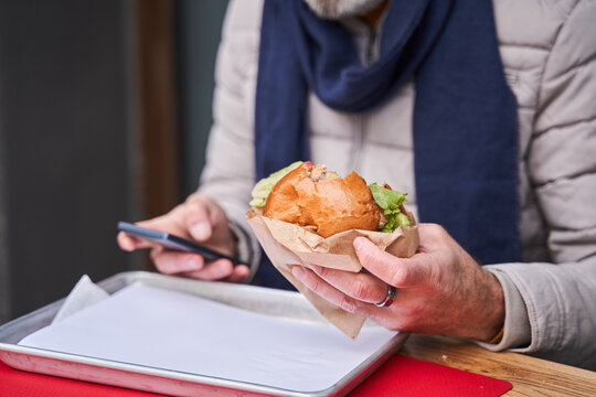 Caucasian senior man holding smartphone and tasty burger at the both hand while eating
