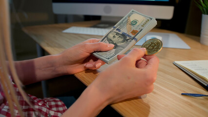 Crop view of female hands holding metal bitcoin and bundle of dollar banknotes sitting at wooden desk with notepad and computer monitor at night