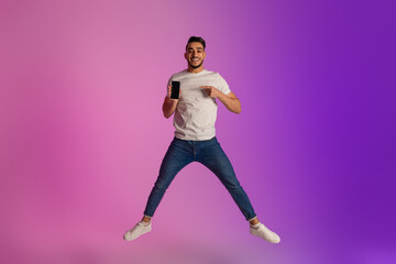 Fototapeta na wymiar Handsome young Arab guy jumping and showing smartphone with blank screen in neon light, mockup