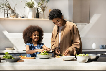 Happy African American young mom and daughter girl preparing dinner together, slicing fresh...