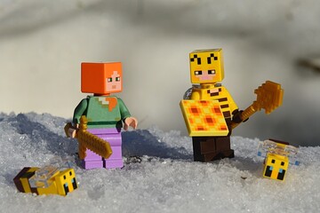 Obraz premium LEGO Minecraft figures of main character Alex with golden sword and Beekeeper with honeycomb and golden shovel, surrounded by snow and LEGO bees, winter afternoon sunshine. 