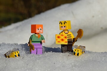 Obraz premium LEGO Minecraft figures of main character Alex and Beekeeper with honeycomb and golden shovel, surrounded by snow and LEGO bees, winter afternoon sunshine. 