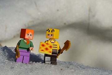 Obraz premium LEGO Minecraft Alex with golden sword and Beekeeper figure with golden shovel and honeycomb talking in front of snow cave, daylight sunshine. 