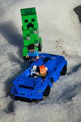 Obraz premium LEGO Minecraft Alex rescuing Steve from attack of giant green explosive Creeper mob, lifting him on board of LEGO Creator McLaren Elva light super sport car in snowy landscape. 