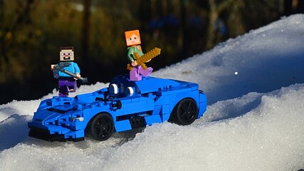 Obraz premium LEGO Minecraft figures of Alex with golden sword and Steve with iron pickaxe standing on LEGO model of McLaren Elva, blue light supersport car, stuck in snow.
