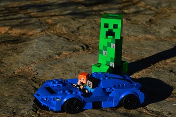 Obraz premium LEGO Minecraft figures of Steve and Alex escaping from dangerous explosive giant Creeper mob on light supersport car McLaren Elva, blue color, on rough stony road.