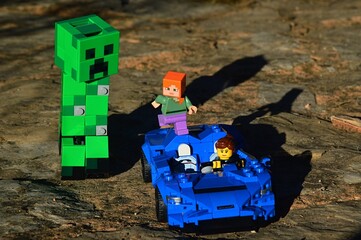 Obraz premium LEGO Minecraft Alex and racer girl from LEGO Creator series escaping on light supersport car McLaren Elva from giant green explosive Creeper mob on rough stony road. Daylight sunshine. 