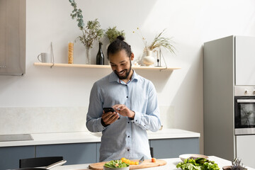 Happy hipster Black chef man using mobile phone in home kitchen, cooking salad, chopping fresh...