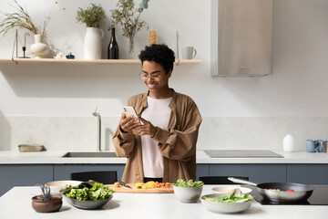 Happy Black food blogger girl taking picture of salad ingredients on mobile phones, recording video, shooting cooking process, reading recipe on Internet, chatting online in home kitchen