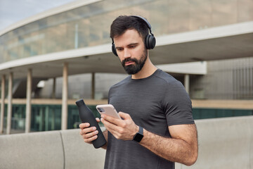 Muscular athletic man looks at smartphone screen downloads songs in playlist for training holds...