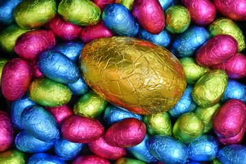 Pile or group of multi colored and different sizes of colourful foil wrapped chocolate easter eggs in pink, blue, yellow and lime green with a large gold egg in the middle.