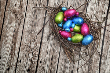Foil wrapped colourful easter eggs in pink, green, blue and yellow in a natural nest made of sticks and twigs, against a multi grain brown white grey wooden background.