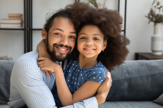 Happy Afro American dad hugging cute curly haired pre teen daughter kid with cheek to cheek touch, looking at camera, smiling. Father embracing kid with love, affection, gratitude. Head shot portrait