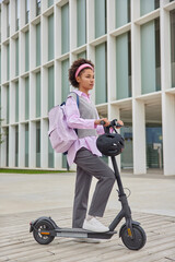Full length shot of curly haired woman in casual clothes carries rucksack rides electric scooter in...