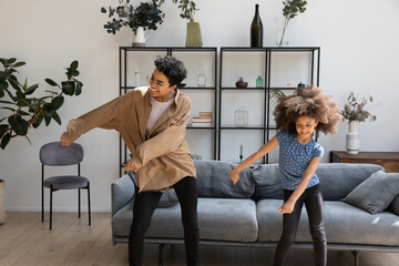 Excited funny Black mom and curly haired daughter kid teaching dance to music in living room, enjoying activity, exercising at home, laughing. Elder and younger sisters having fun at home