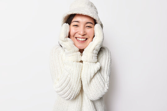 Happy Asian woman has tender look healthy skin touches face gently smiles joyfully has good mood enjoys good winter weather wears hat mittens and knitted sweater isolated over white background.