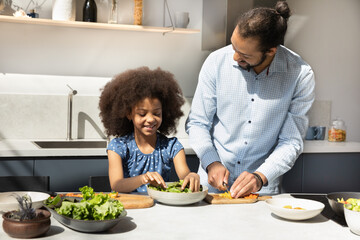 Happy millennial African father and cute 7s daughter girl preparing fresh vegetable salad in home kitchen, talking, chatting, laughing. Dad teaching kid to cook healthy meal from organic food