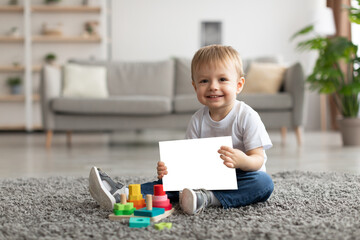 Portrait of adorable little child boy holding blank card and smiling at camera, sitting on floor carpet. Mockup image