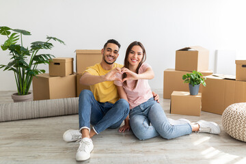 Loving young international couple making heart with their hands, sitting on floor among boxes,...
