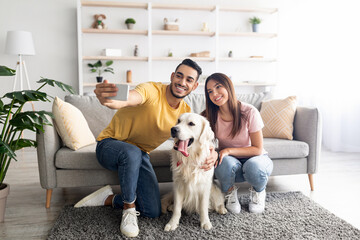 Positive young diverse couple taking selfie with their dog while sitting on floor at home, full length
