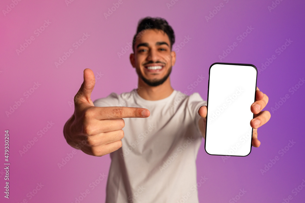 Poster smiling young arab guy pointing at cellphone with mockup for mobile app on screen in neon light, sel