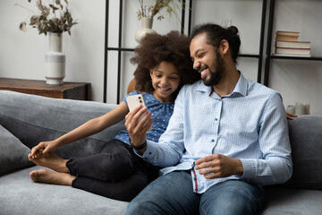 Fototapeta Happy Afro American dad and gen Z teen daughter kid using smartphone at home together, resting on comfortable sofa in living room, making video call on mobile phone, shopping online, browsing internet obraz