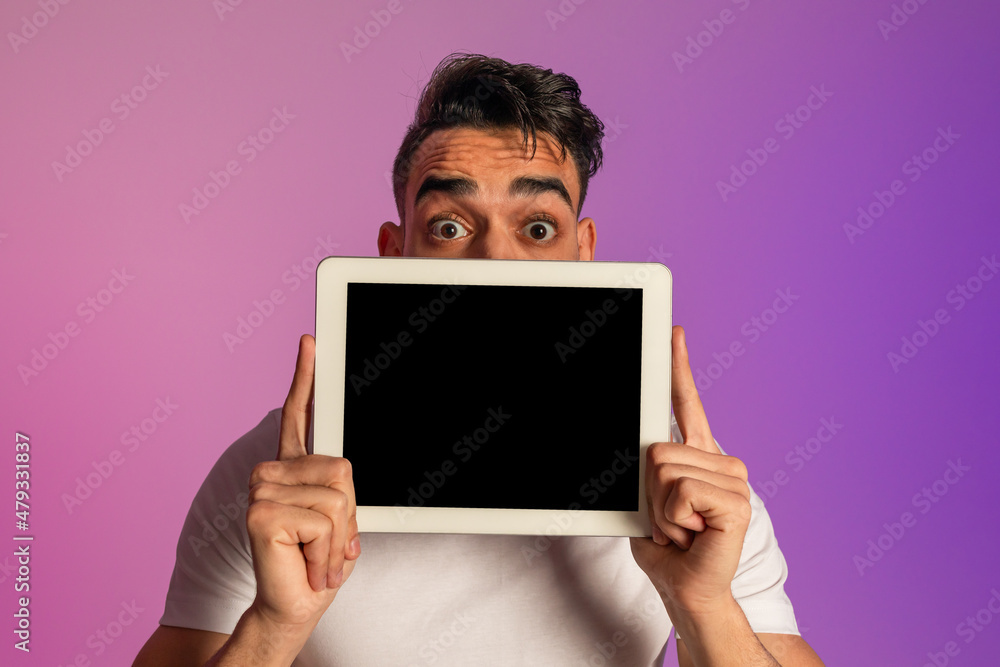 Poster shocked millennial arab man hiding behind tablet with blank screen in neon light, mockup for app or 
