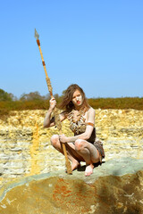 A young woman is dressed as Stone Age warrior. 
Her body and face is covered with mud and dirt.
She is seen posing in a stone quarry.
